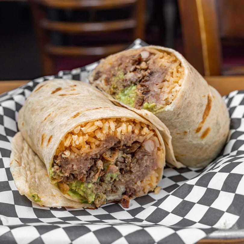 Two La Sureña Taquerias burritos, cut in half, revealing rice, meat, and guacamole on checkered paper.