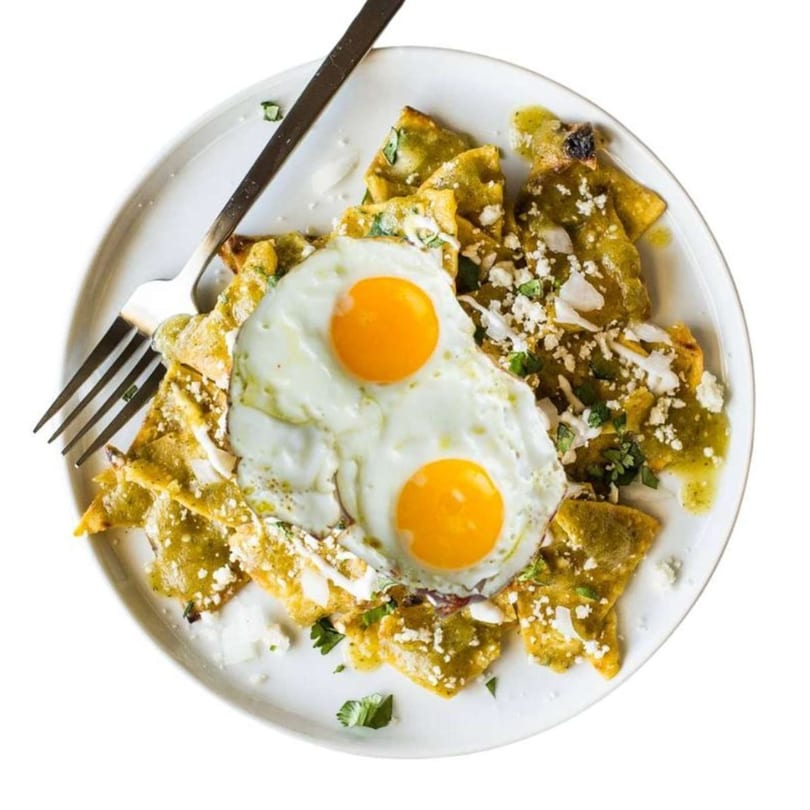Chilaquiles Verdes with two fried eggs, crumbled cheese, and cilantro on a white plate with a fork.
