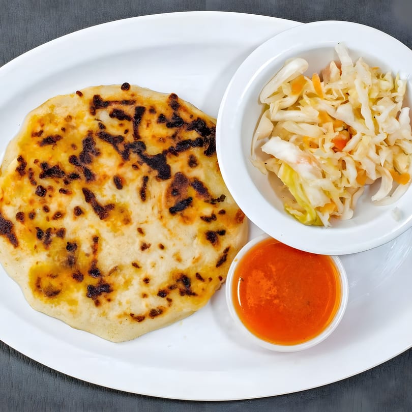 A golden-brown pupusa with charred spots, served with curtido and red salsa on a white plate.